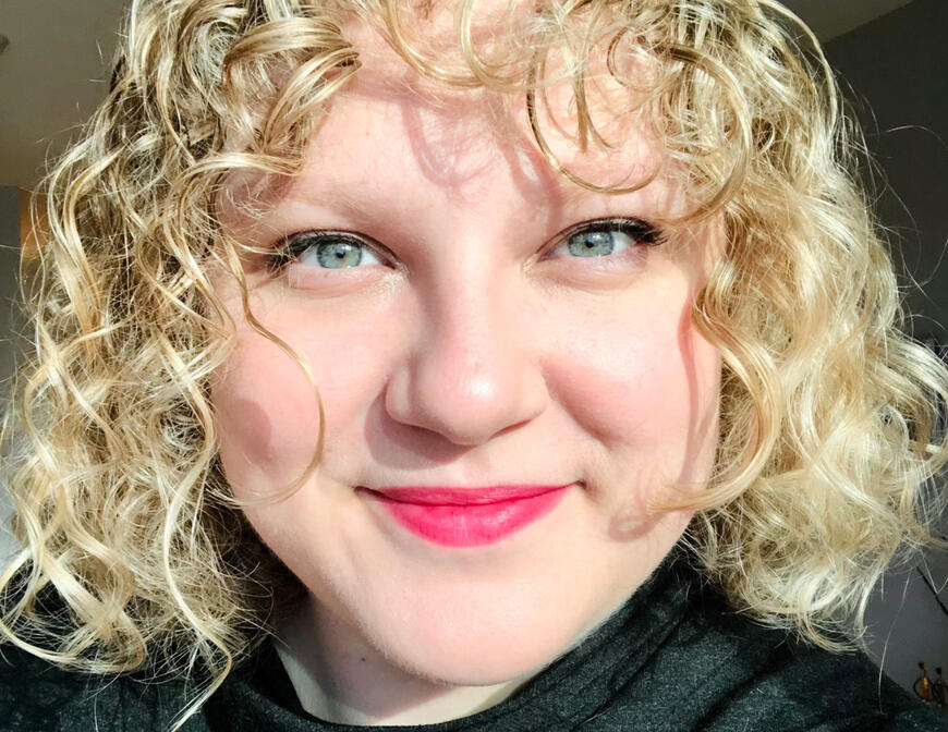 A selfie of Nat. They look directly into the camera, smiling. She is white, with blonde hair, blue eyes, and wears red lipstick.