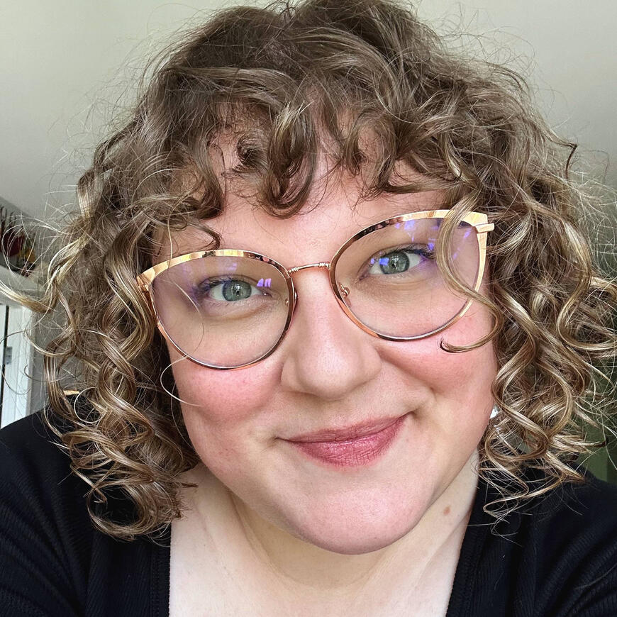 A selfie of Nat. They look directly into the camera, smiling. She is white, with blonde hair, blue eyes, and wears red lipstick.